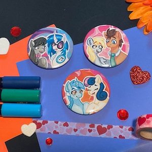 May include: Three round buttons with colorful cartoon images of ponies. The first button features two ponies, one with a blue mane and a blue and white striped shirt, the second pony has a gray mane and a pink shirt. The second button features two ponies, one with a pink mane and a blue shirt, the second pony has a yellow mane and a blue shirt. The third button features two ponies, one with a blue mane and a blue shirt, the second pony has a pink mane and a pink shirt.