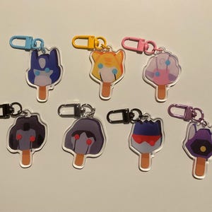 May include: Eight colorful character keychains shaped like popsicles with different colored eyes and expressions. The keychains are attached to silver, gold, and purple key rings.