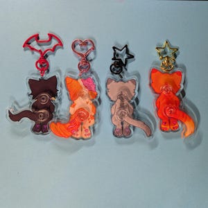 May include: Four acrylic keychains featuring cartoon cat figures viewed from behind. The first has a red bat-shaped clasp, the second a rose gold heart clasp, the third a black star clasp, and the fourth a gold star clasp with orange fur.