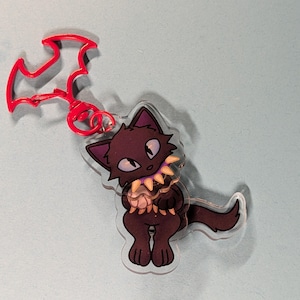 May include: A clear acrylic keyring featuring a dark brown cartoon cat with large eyes and a purple collar with yellow spikes. The cat is attached to a bright red metal clasp shaped like a bat silhouette, providing a secure way to hang the charm.