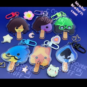 May include: A collection of six acrylic keychains shaped like popsicles, each featuring a unique character design. The set includes various colored clasps, small charms like a star, moon, and heart, and text reading Only on Etsy! Not on Etsy? It's a scam!
