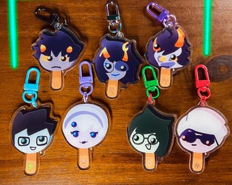 May include: A selection of cartoon character keyrings in various designs. Each keyring features a unique character design on a popsicle stick shape, with a metal clasp for attachment. The keyrings are in various colours, including purple, blue, and green.