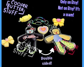 May include: A double-sided acrylic keychain featuring two cartoon figures, one wearing a black shirt with the Roblox logo and a red cap, the other in a blue shirt and green pants. Includes yellow bow charms, a candy charm, and a heart charm.