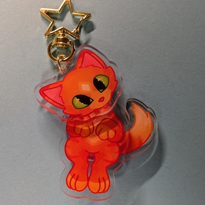 May include: An acrylic keyring featuring a cartoon orange tabby cat with large green eyes and a long tail. The cat is depicted in a standing position with its front paws tucked near its chest. It is attached to a gold-coloured star-shaped clasp.