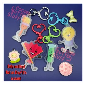 May include: A collection of acrylic character keychains featuring a paintbrush, a red fan, a green test tube, and a lightbulb, each with a heart-shaped clasp. Includes small charms of a flower and a pink cookie. Text reads COOPER BUTTER STUFF.