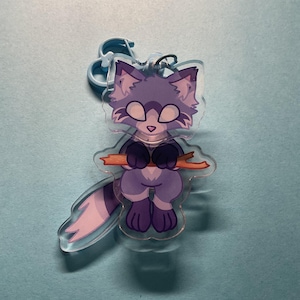 May include: A clear acrylic keyring featuring a cartoon raccoon character with purple and light lavender fur. The raccoon has large white eyes, dark purple paws, and a long striped tail, and is shown holding a small orange branch in its hands.