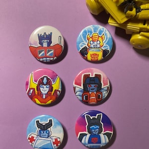 May include: Six round pinback buttons featuring different Transformers characters. The buttons are colorful and feature a variety of characters, including Optimus Prime, Bumblebee, Starscream, and others.