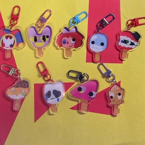 Puede incluir: Una colecci&oacute;n de nueve llaveros de acr&iacute;lico con forma de diversos helados de palo con caras de personajes coloridos. Cada llavero cuenta con un cierre de mosquet&oacute;n de metal en colores como rojo, azul, plateado y rosa, unido a un dije de acr&iacute;lico.