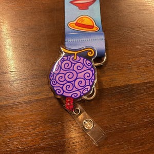 May include: A lanyard with a blue strap featuring cartoon images of a bowl and a straw hat. A purple, swirl-patterned fruit-shaped badge reel is attached. The reel has a clear plastic strap and a metal clip.