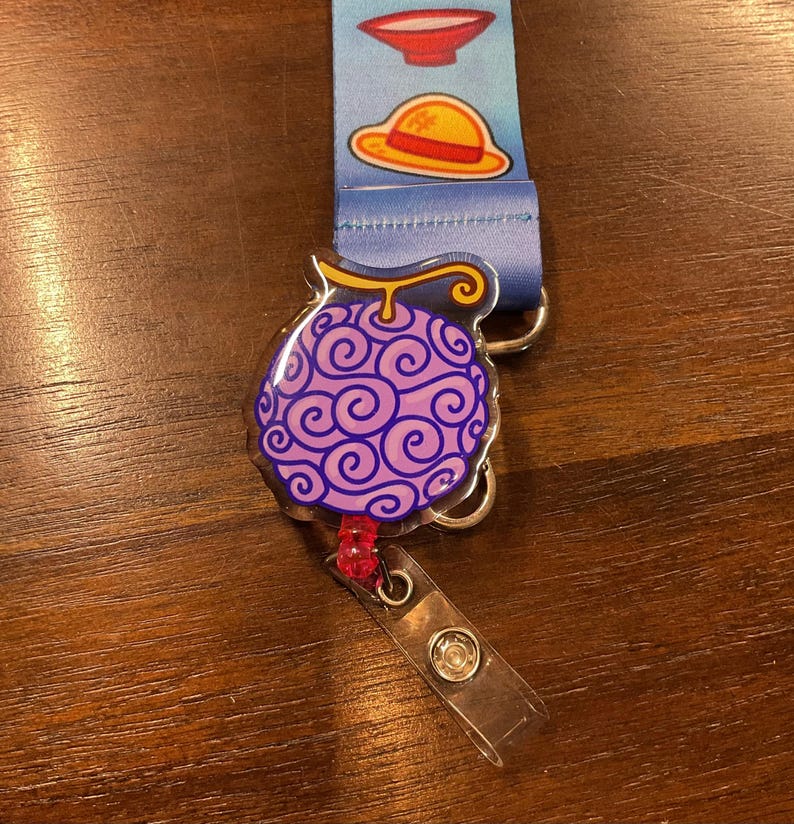 May include: A lanyard with a blue strap featuring cartoon images of a bowl and a straw hat. A purple, swirl-patterned fruit-shaped badge reel is attached. The reel has a clear plastic strap and a metal clip.