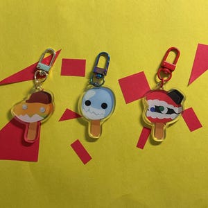Puede incluir: Tres dijes de acr&iacute;lico con forma de paletas heladas y caras de dibujos animados. El dije izquierdo tiene una gorra marr&oacute;n, el central es azul claro con un borde dentado y el derecho es rojo y blanco con una parte superior negra.