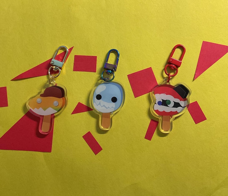 Puede incluir: Tres dijes de acr&iacute;lico con forma de paletas heladas y caras de dibujos animados. El dije izquierdo tiene una gorra marr&oacute;n, el central es azul claro con un borde dentado y el derecho es rojo y blanco con una parte superior negra.