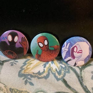 May include: Three round buttons featuring different Spider-Man characters. The first button has a black Spider-Man with a purple background. The second button has a red Spider-Man with a green background. The third button has a white Spider-Man with a blue background.