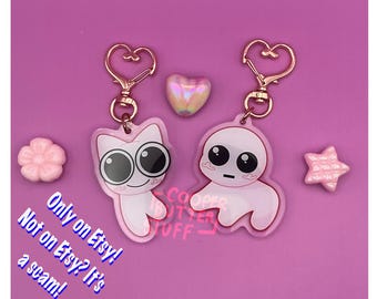 May include: Two pink acrylic keychains with rose gold heart-shaped clasps. One features a cat-like character with large black eyes, the other a round-headed figure. Includes a small flower, star, and heart charm. Text reads: Only on Etsy!