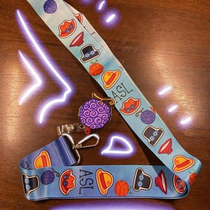 May include: A light blue lanyard with cartoon illustrations of hats, berries, and fruit. The lanyard has a metal clasp and the letters "ASL" printed on it. A purple and pink pin with a fruit design is also visible.