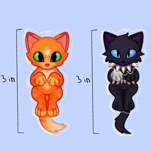 May include: Two acrylic charms, each measuring 7.62 cm in height. One charm features an orange tabby cat with green eyes, while the other depicts a black cat with blue eyes and a spiked collar. Both charms have a clear outline and a hole at the top.