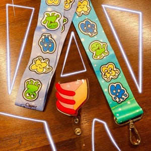 May include: Two lanyards with cartoon character designs in blue and teal, and a clear badge reel with a cake slice design. The lanyards feature repeating patterns of various cartoon characters. One lanyard is light blue, the other is teal.