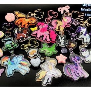 May include: A collection of acrylic keychains featuring various cartoon pony and animal characters, including a blue pegasus with a rainbow mane, a pink pony, and a purple unicorn. The set includes heart, star, and butterfly charms with metal clasps.