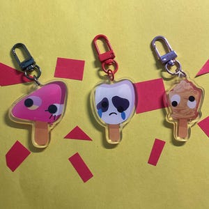 Puede incluir: Tres dijes de acr&iacute;lico con forma de paletas de helado y ojos m&oacute;viles. El primero es rosa con un lado rosa oscuro, el segundo es blanco con una cara triste y l&aacute;grimas azules, y el tercero es color canela con textura arremolinada y gancho.
