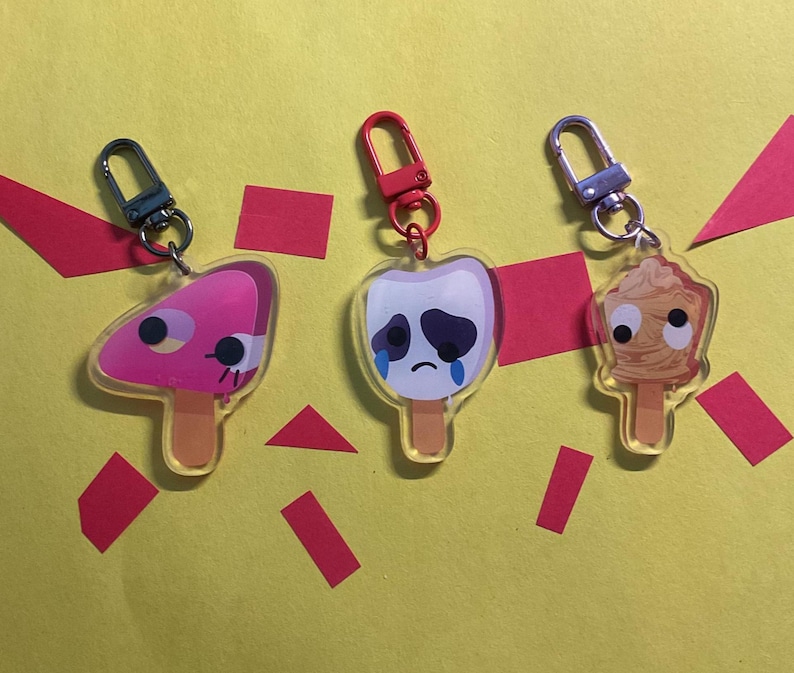Puede incluir: Tres dijes de acr&iacute;lico con forma de paletas de helado y ojos m&oacute;viles. El primero es rosa con un lado rosa oscuro, el segundo es blanco con una cara triste y l&aacute;grimas azules, y el tercero es color canela con textura arremolinada y gancho.