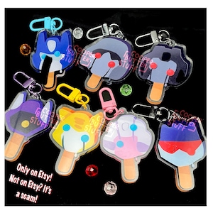 May include: Seven acrylic keychains shaped like popsicles with various character designs, each featuring a metal lobster clasp. The designs include blue, purple, yellow, and pink color schemes with wooden-style sticks. Text reads: Only on Etsy! Not on Etsy? It's a scam!