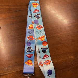 May include: A light blue lanyard with a repeating pattern of cartoon illustrations and the letters "ASL". The lanyard has a metal clasp and a matching blue fabric. The illustrations include hats, berries, and other whimsical designs.