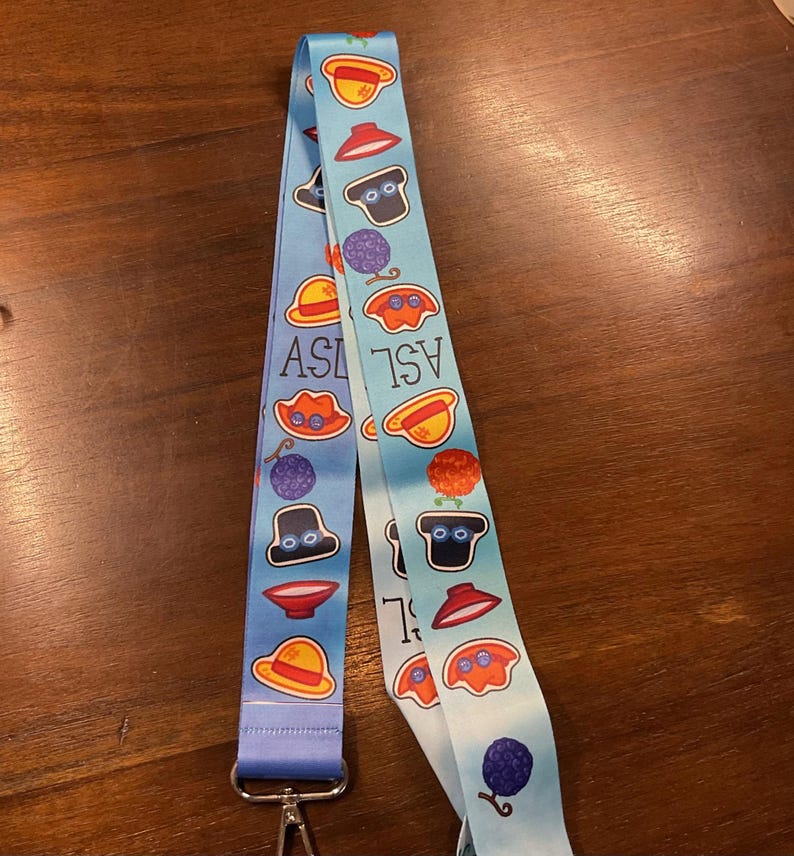 May include: A light blue lanyard with a repeating pattern of cartoon illustrations and the letters "ASL". The lanyard has a metal clasp and a matching blue fabric. The illustrations include hats, berries, and other whimsical designs.