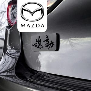Può includere: Un emblema rettangolare nero con caratteri kanji giapponesi fissato sul bagagliaio grigio di un veicolo Mazda. L'emblema è posizionato sotto il logo Mazda, visualizzato in un riquadro bianco sopra il gruppo ottico posteriore dell'auto.