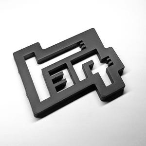 May include: A black, three-dimensional, geometric object featuring a cutout design that spells out the word "LEFT" in a blocky, sans-serif font. The object has a matte finish and sharp, angular edges, creating a distinct, stencil-like appearance.