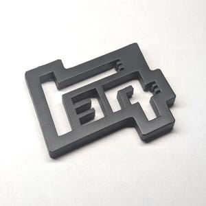May include: A matte dark gray, flat, geometric object featuring a cutout design that forms the letters "EQ". The shape has a rectangular outer frame with internal angular lines and a stepped, blocky silhouette with sharp, clean edges.