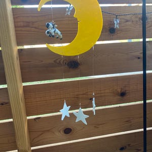 Könnte beinhalten: Ein gelber Halbmond-Mobile mit einer weißen Kuhfigur, die daran hängt. Drei weiße Sterne hängen am Mond, jeder an einer separaten Schnur.