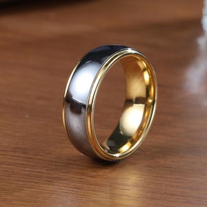 Puede incluir: Un anillo de carburo de tungsteno negro y dorado con un borde biselado.