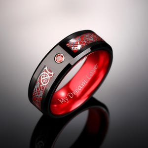 以下が含まれることがあります： 赤い内側バンドと銀色のケルト結び目のデザインが施された、黒色のタングステンカーバイド製のリング。リングには小さな赤い宝石と、内側に「My Dream Love」という刻印があります。