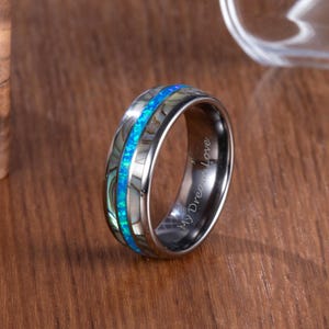 May include: A silver ring with a blue opal inlay and mother-of-pearl accents. The ring has a polished finish and the words "My Dream Love" are engraved on the side. The ring is displayed on a wooden surface.