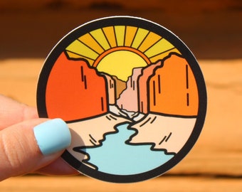 May include: A round sticker with a colorful illustration of a canyon with a river running through it. The sun is setting behind the canyon, casting a warm glow on the scene.