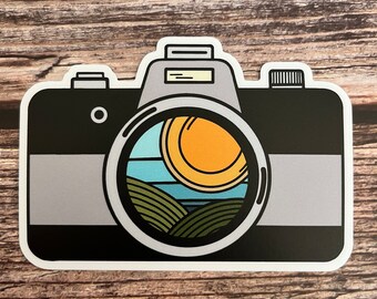May include: A black and gray camera sticker with a viewfinder showing a cartoon landscape with a sun, blue sky, and green hills.