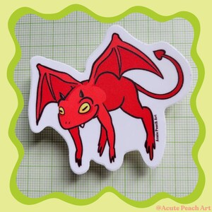 May include: A die-cut sticker featuring a cartoon illustration of a bright red winged creature with yellow eyes, small horns, and a pointed tail. The design includes the text Acute Peach Art printed in small black letters on the right side.
