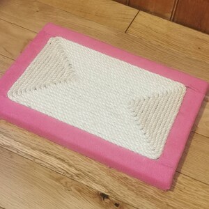Puede incluir: Un rascador de gato rectangular de color rosa con una superficie de cuerda blanca.