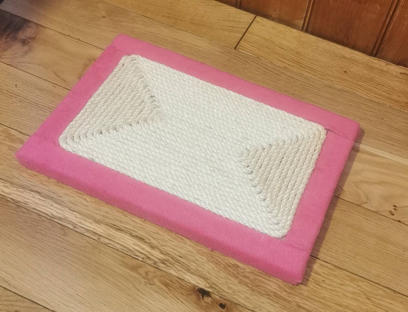 Puede incluir: Un rascador de gato rectangular de color rosa con una superficie de cuerda blanca.