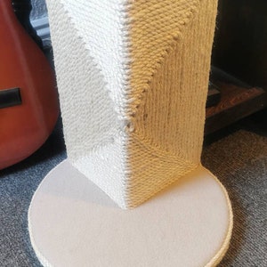 May include: A beige cat scratching post with a round base and a square top covered in sisal rope. The post is made of natural materials and is designed to help cats scratch and play.