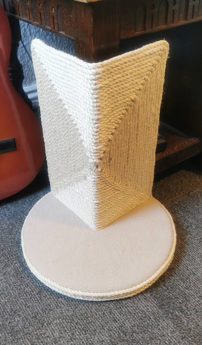 May include: A beige cat scratching post with a round base and a square top covered in sisal rope. The post is made of natural materials and is designed to help cats scratch and play.