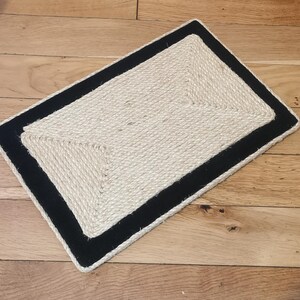 Puede incluir: Una alfombra rectangular de fibra natural con borde negro. La alfombra est&aacute; hecha de cuerda trenzada y tiene un patr&oacute;n geom&eacute;trico.
