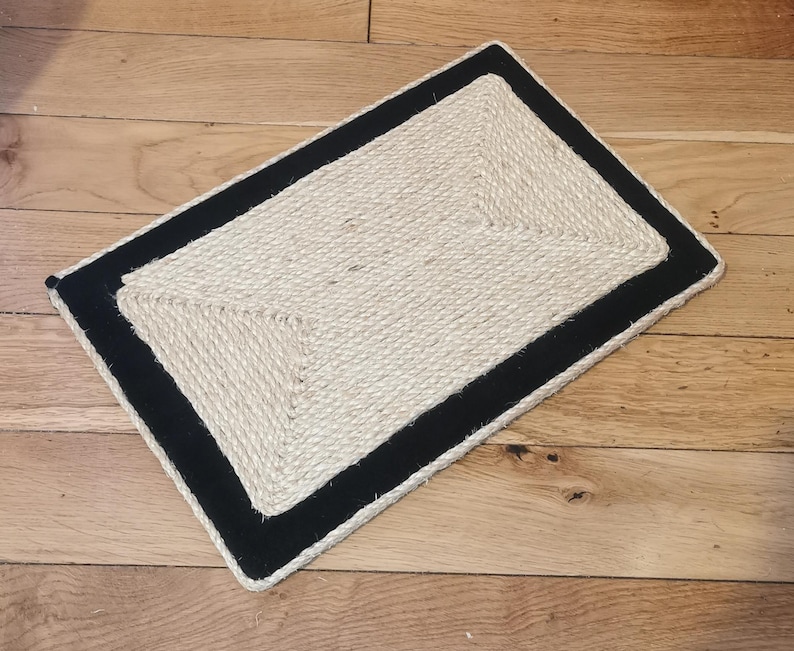 Puede incluir: Una alfombra rectangular de fibra natural con borde negro. La alfombra est&aacute; hecha de cuerda trenzada y tiene un patr&oacute;n geom&eacute;trico.