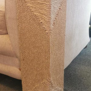 May include: A close-up of a beige sofa armrest with a tan, rope-like corner protector. The protector is made of woven rope and is designed to protect the furniture from scratches.