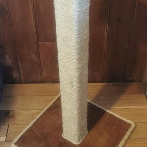 May include: A tall, natural sisal-wrapped cat scratching post with a square wooden base and top. The post is on a wood floor.