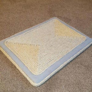 Puede incluir: Un rascador para gatos azul claro con una superficie de sisal beige. El rascador es rectangular y tiene una secci&oacute;n central elevada.