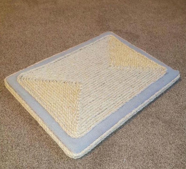 Puede incluir: Un rascador para gatos azul claro con una superficie de sisal beige. El rascador es rectangular y tiene una secci&oacute;n central elevada.