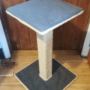 May include: A square cat scratching post with a grey, fabric-covered platform and a sisal rope-covered post. The post is on a wooden floor.