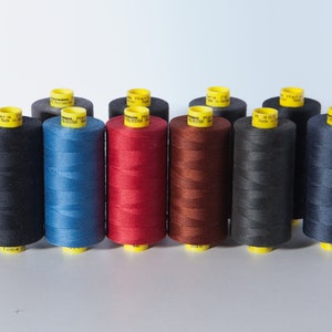 Puede incluir: Una colección de diez carretes de hilo en varios colores, incluyendo negro, azul, rojo, marrón y gris. Cada carrete tiene una etiqueta amarilla con el texto "Gütermann" y "Made in Germany".