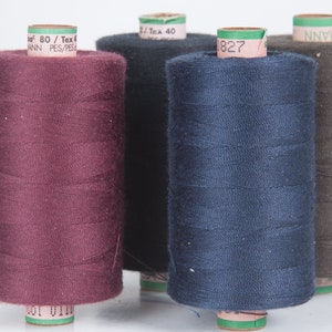 Op de afbeelding: Vier spoelen garen in verschillende kleuren: bordeauxrood, donkerblauw, bruin en grijs. De spoelen zijn gelabeld met de merknaam "MANN" en de tekst "80 / Tex 40".